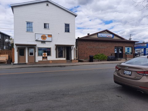 A car is parked on the side of the road in front of a white building with a sign that reads "Pharmacy Choice.".