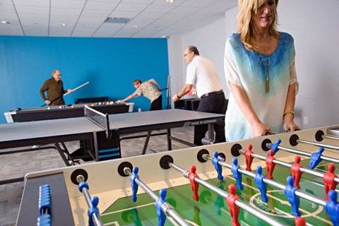 A woman is playing foosball in a room with people playing ping pong in the background.