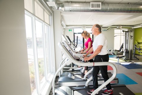 Two people are exercising on a treadmill in a brightly lit gym.