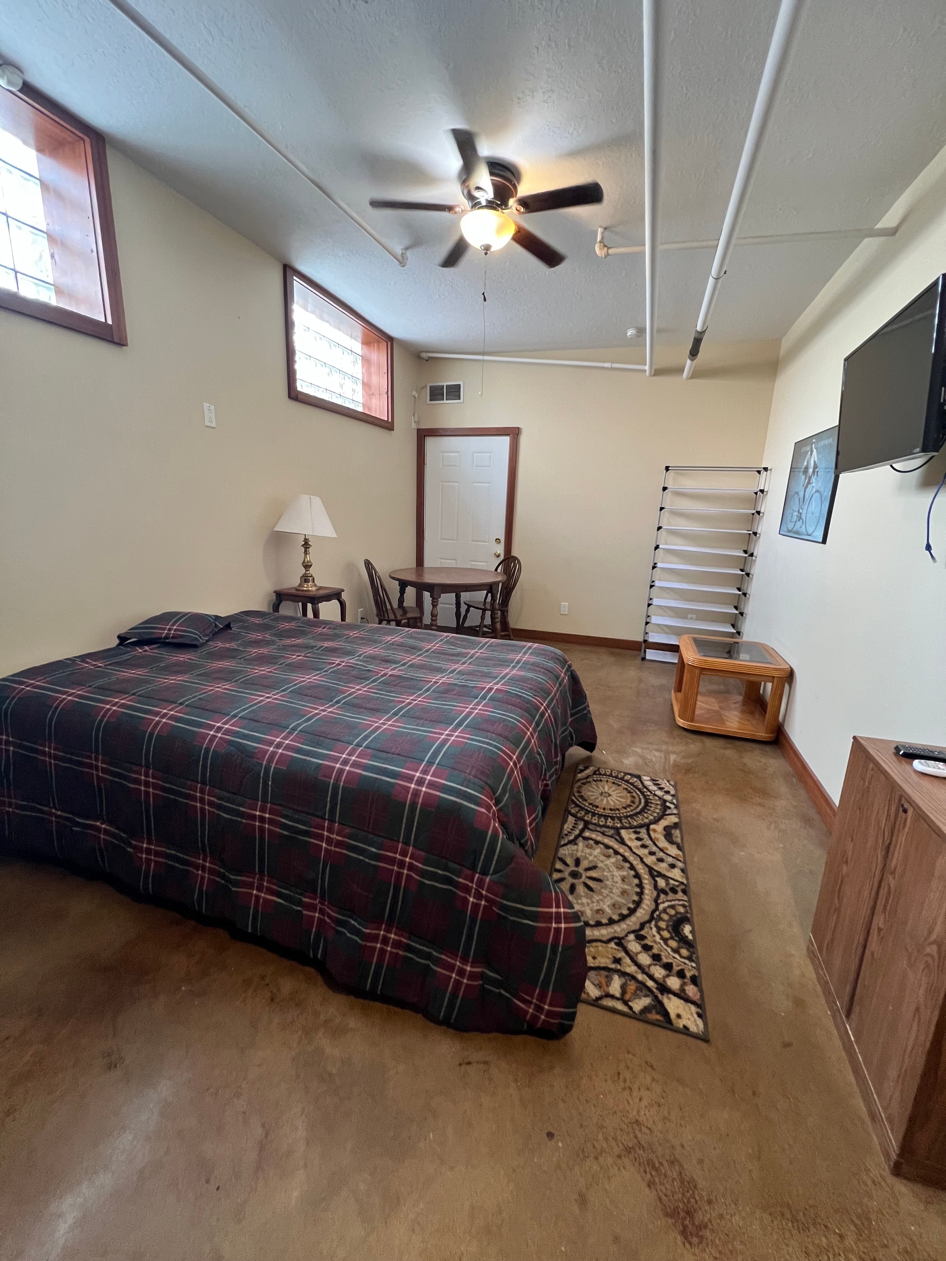 A bedroom with a bed, a chair, a table, a lamp, a ceiling fan, and a wall-mounted TV.