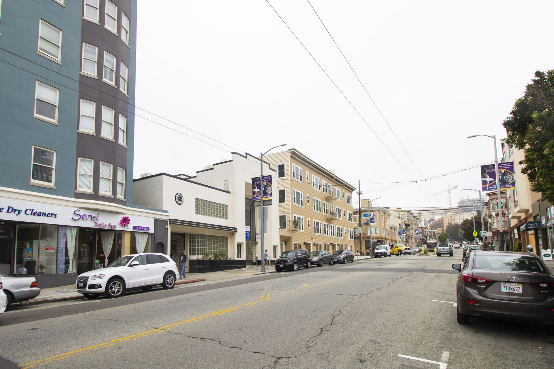A street with cars parked on the side and a dry cleaners storefront.