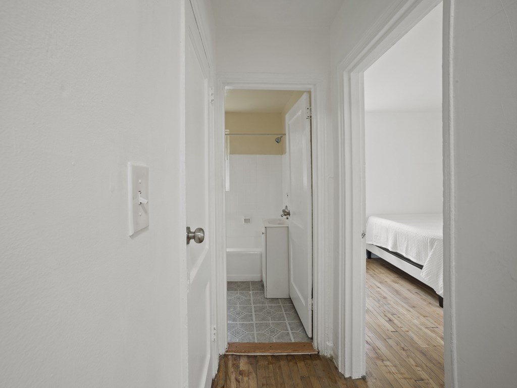 A white hallway with a door on the left and a bed on the right.