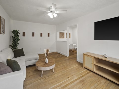 A living room with a white couch, a wooden coffee table, and a TV on the wall.