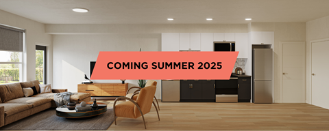 A modern living room with a sofa, coffee table, and a kitchen area in the background, with a sign that says "COMING SUMMER 2025".