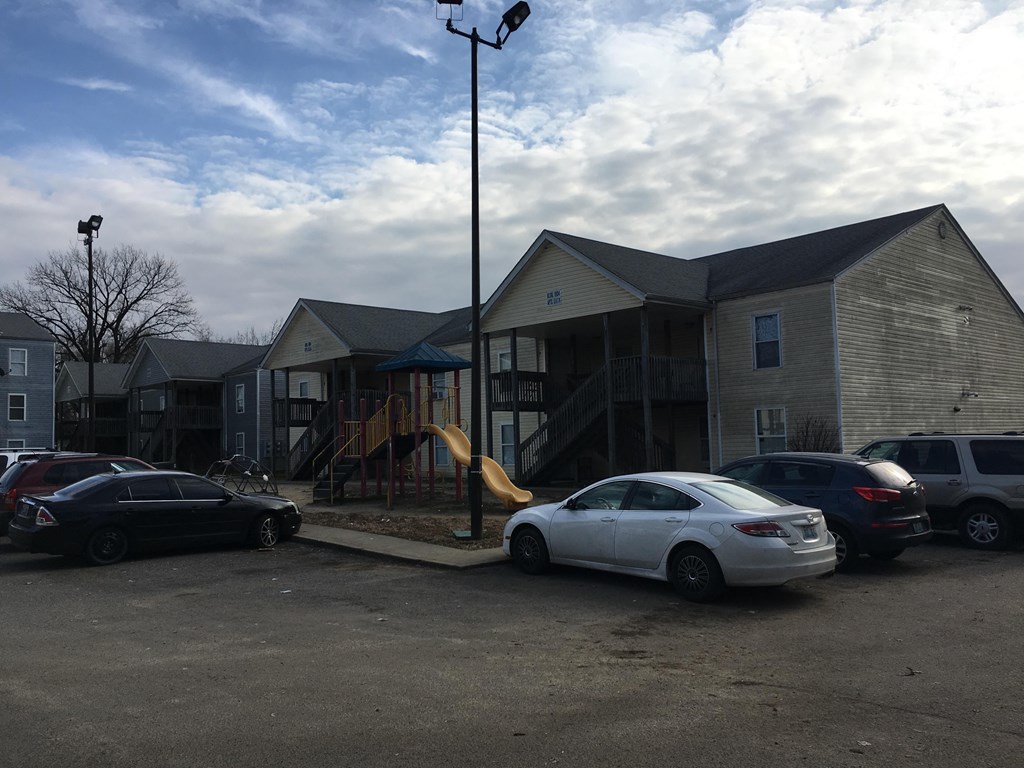 A white car is parked in a lot with other cars and a playground is in the background.