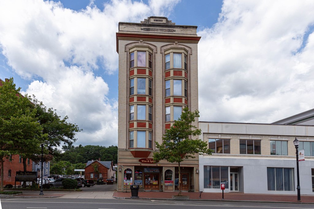 A tall building with a red and white facade stands in front of a white building and trees.
