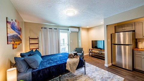 A bedroom with a blue bedspread and a stainless steel refrigerator in the kitchen area.