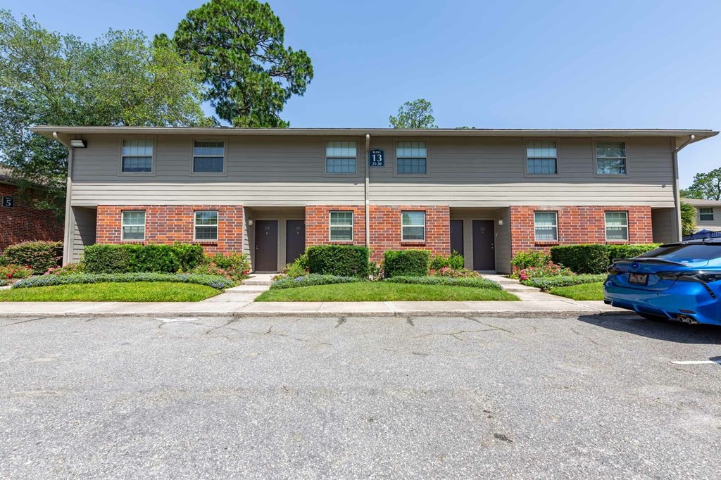 A blue car is parked in front of a two-story building with a brick facade and a grey roof.