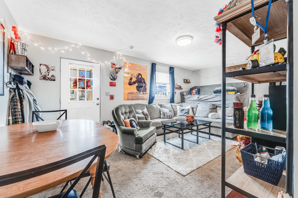 A living room with a grey couch, a coffee table, a dining table, and a shelf with various items.
