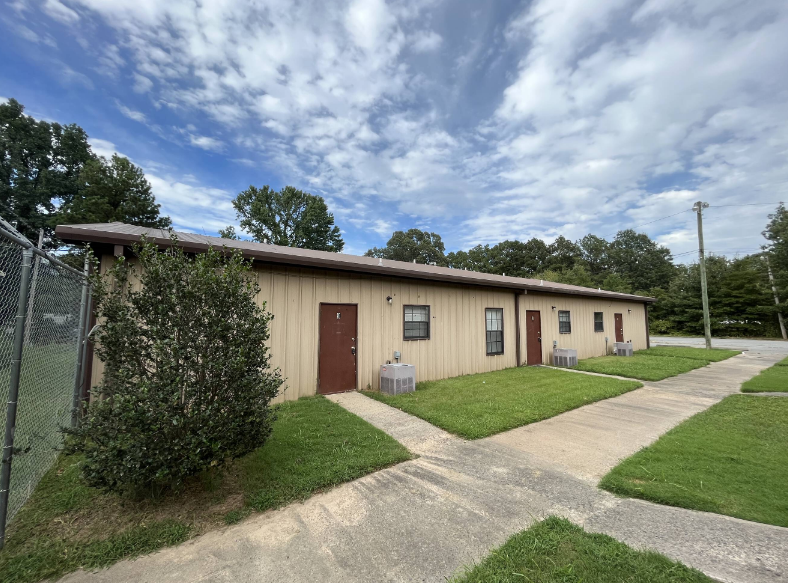 A building with a brown door and windows is surrounded by a grassy area and a fence.