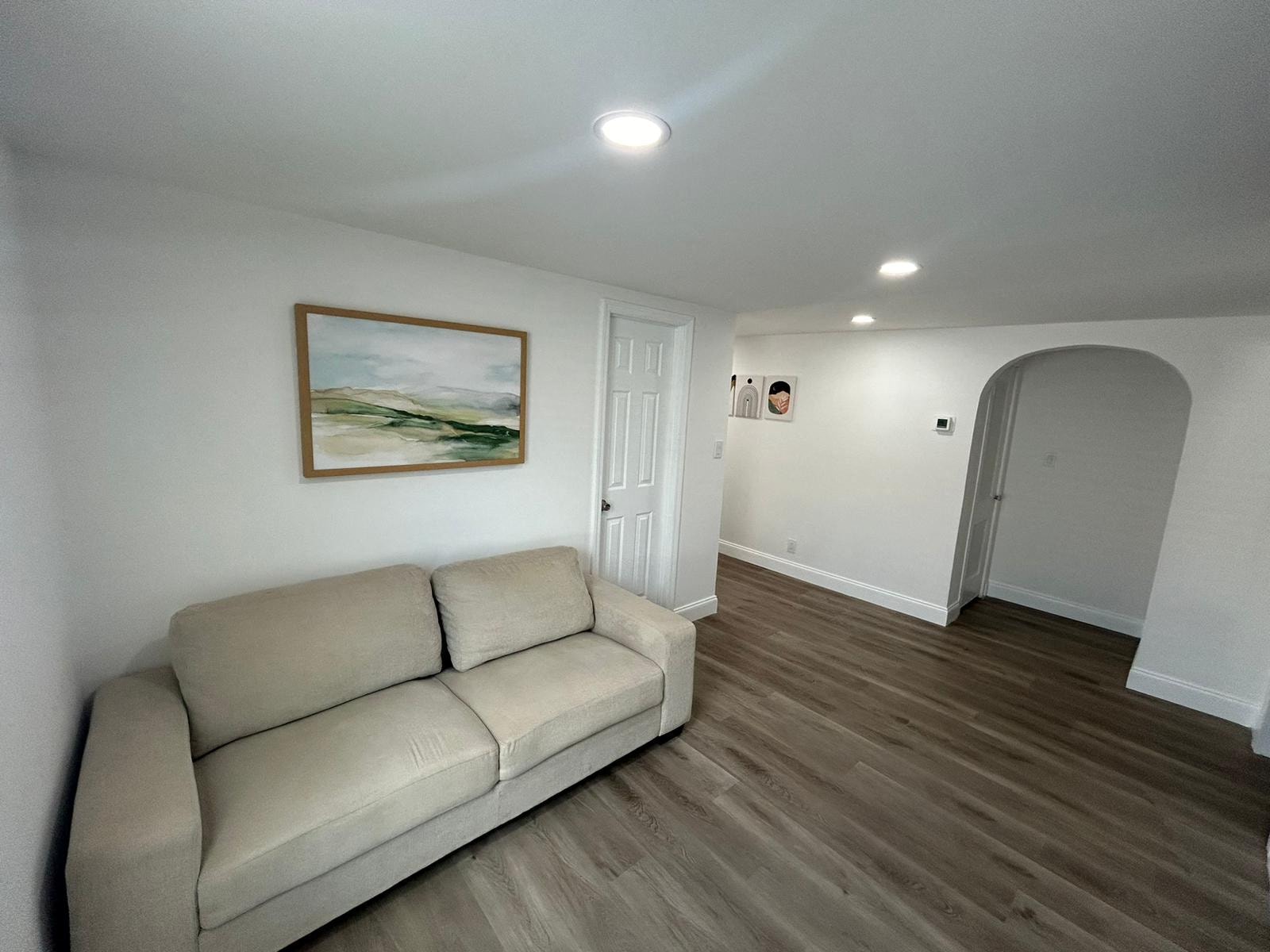 A beige couch sits in a room with a painting on the wall and a white door.