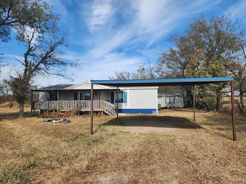 A house with a blue roof and white walls is surrounded by dry grass and trees.