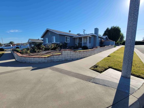A house with a blue roof and a white fence is surrounded by a grassy area and a concrete walkway.