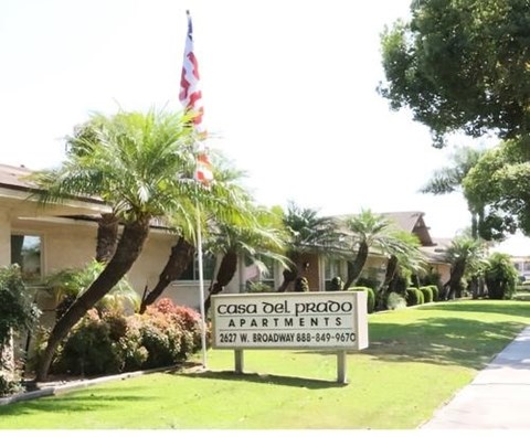 A sign for Coso Del Prado Apartments is in front of a building with trees and an American flag.