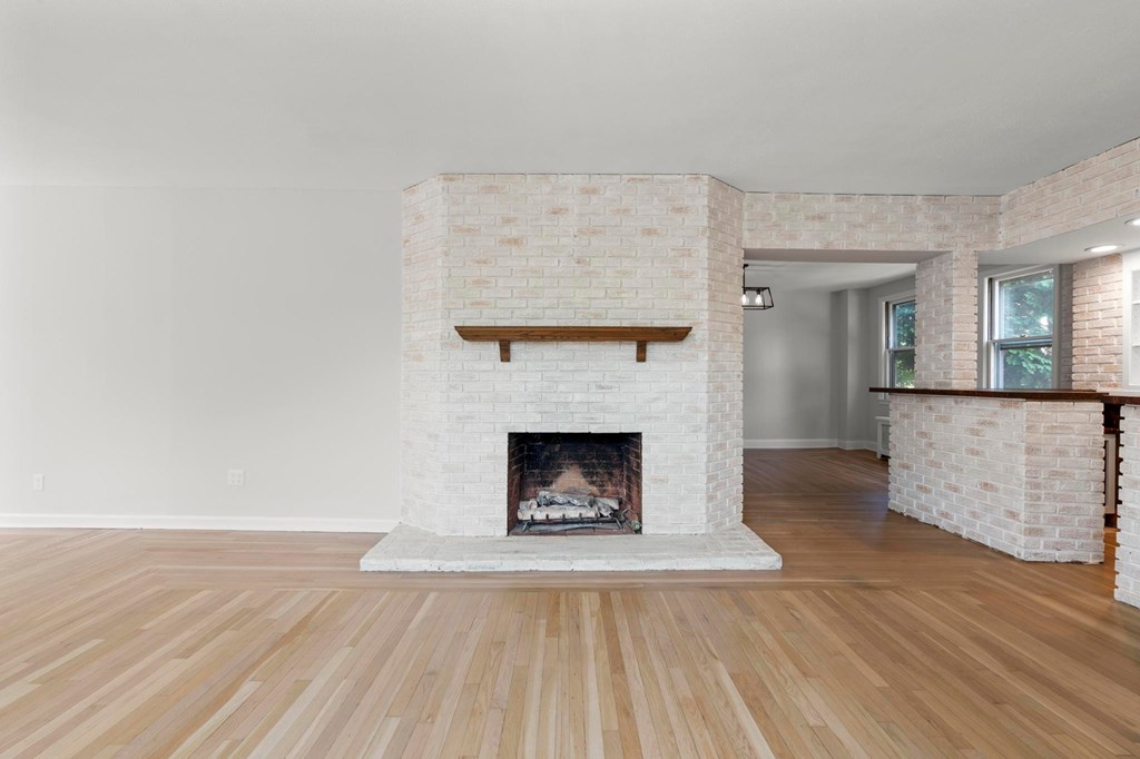 A white brick fireplace with a wooden mantle is in the middle of a room with wooden floors.