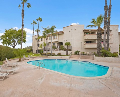 A swimming pool is surrounded by palm trees and lounge chairs in front of a building.