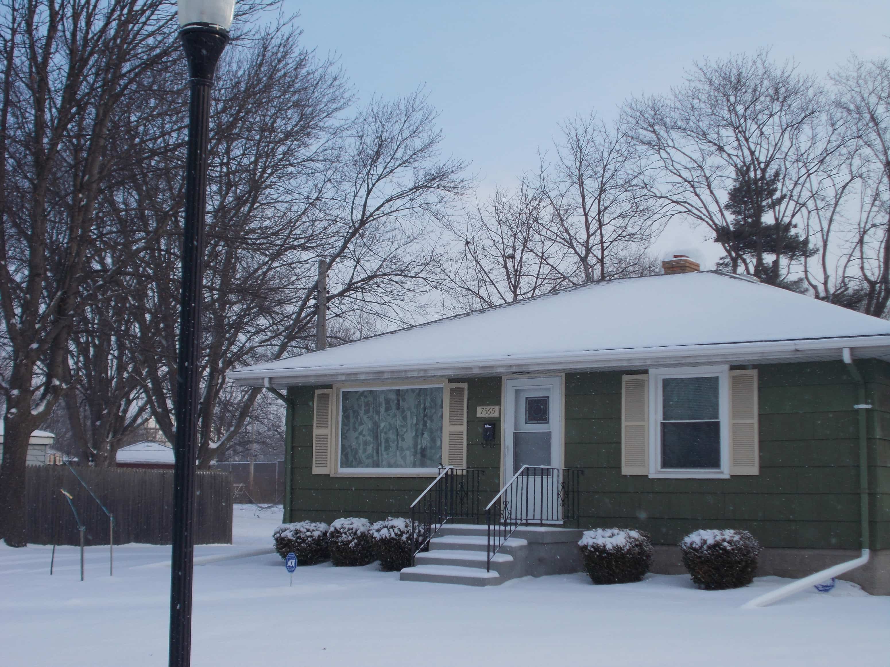 A green house with a white door and windows is surrounded by snow.