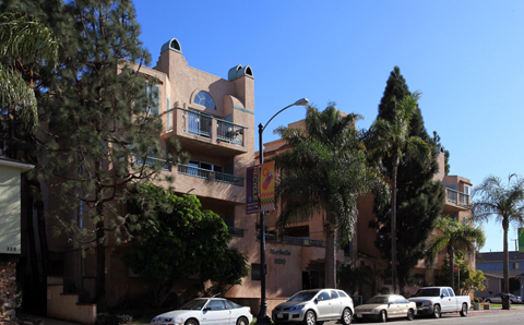 A white car is parked in front of a tan building with a balcony.