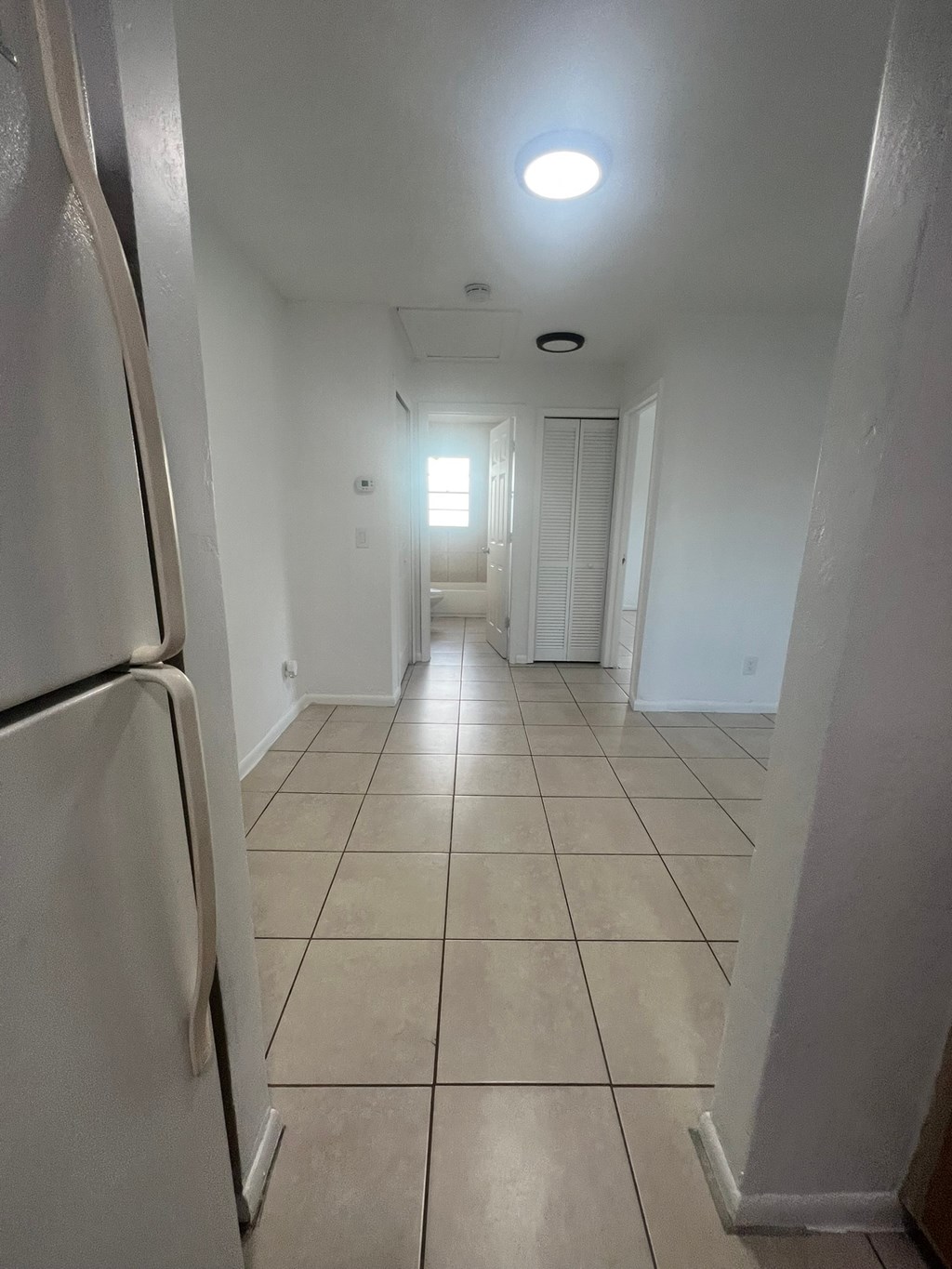 A white refrigerator is on the left side of a narrow hallway with tiled floors and white walls.
