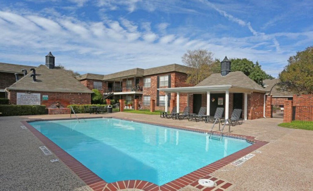 A swimming pool in front of a house with a patio and chairs.