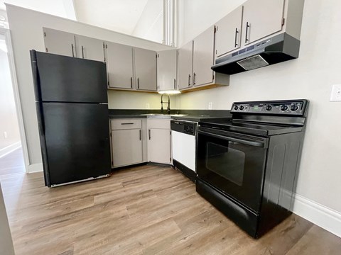 A black refrigerator stands next to a black oven in a kitchen with wooden floors and white walls.