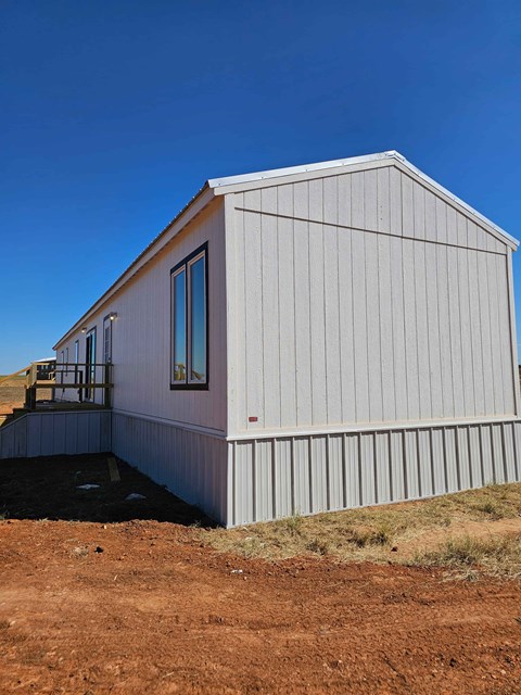 A small white building with a window and a door is situated on a dirt area.