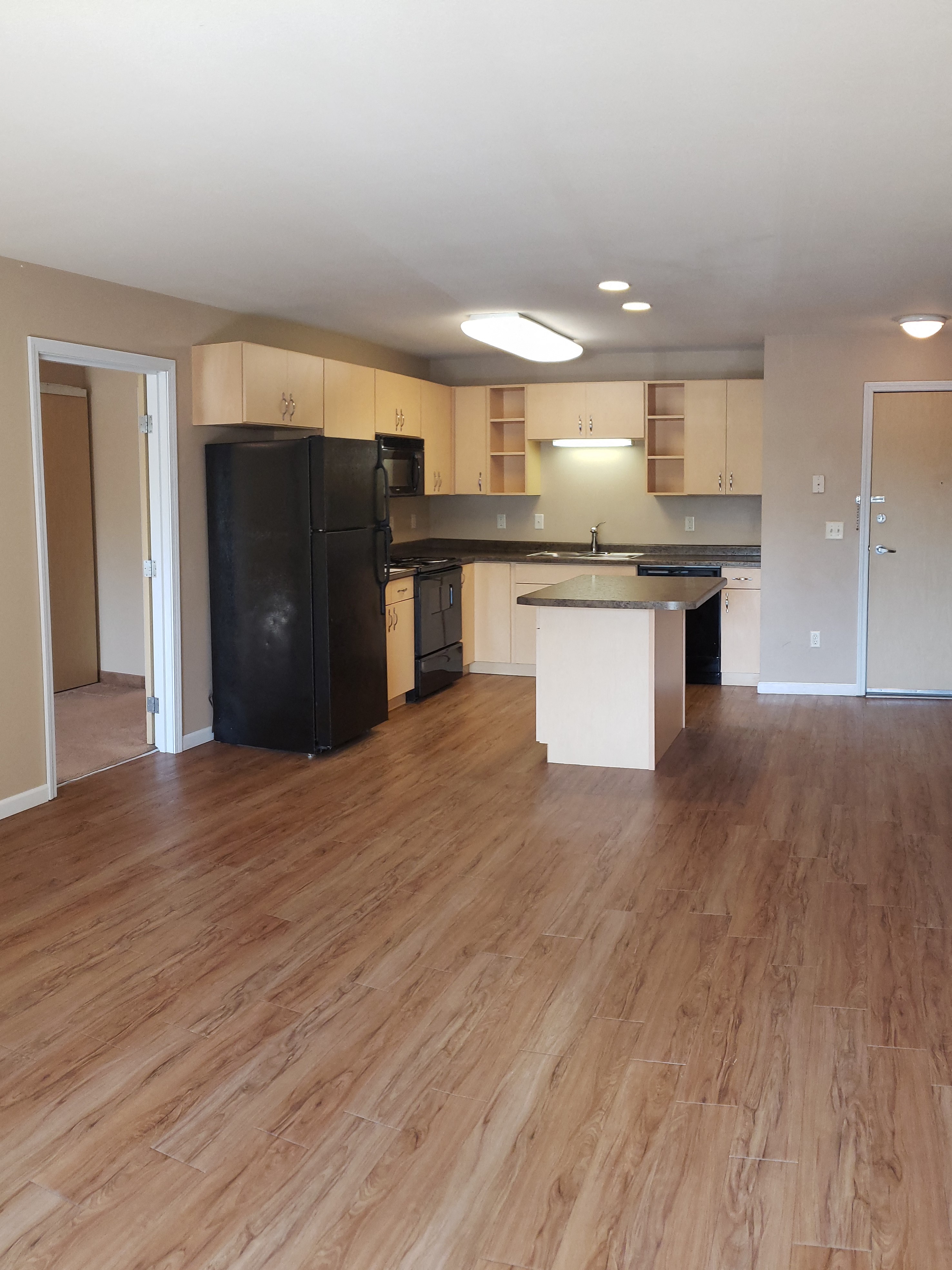 an empty kitchen and living room with wood floors and a black refrigerator