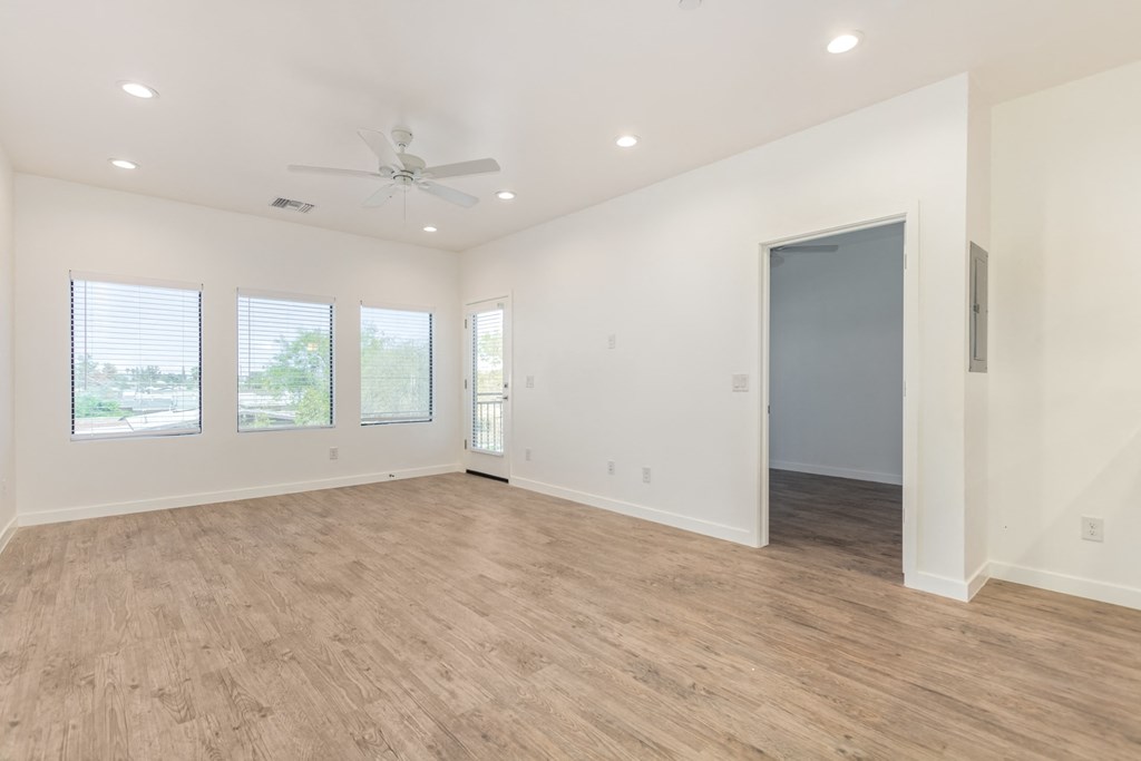 A spacious room with a ceiling fan and lighting, hardwood flooring, and a door on the right side.