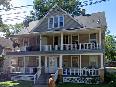 A man stands on the front porch of a two-story house.