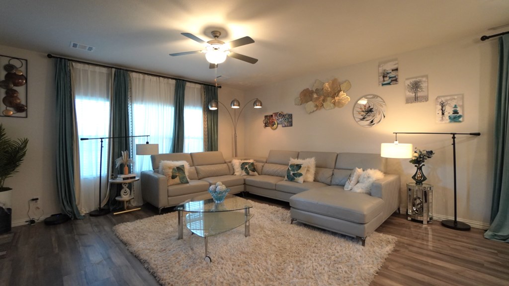 A living room with a white rug, a grey sofa, a glass coffee table, and a ceiling fan.