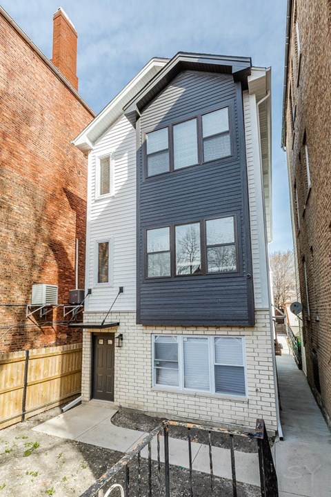 A two-story building with a grey facade stands between a red brick building and a brown wooden fence.