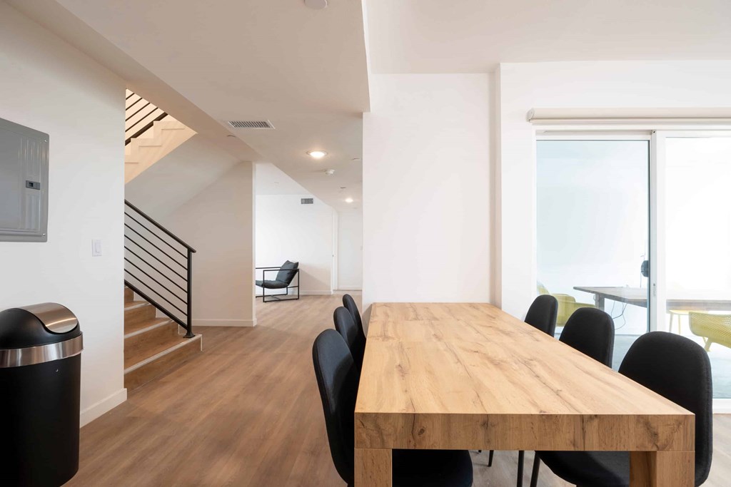 A long wooden table is surrounded by black chairs in a white room with a staircase.