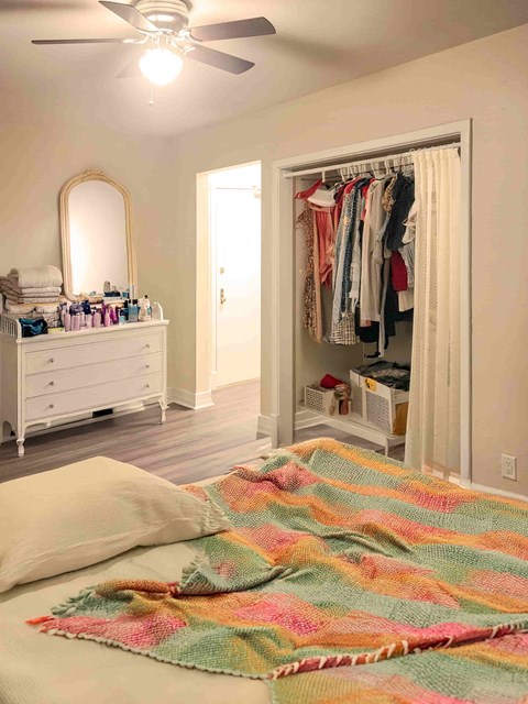 A bedroom with a bed, a dresser, a mirror, a ceiling fan, and a closet with clothes.