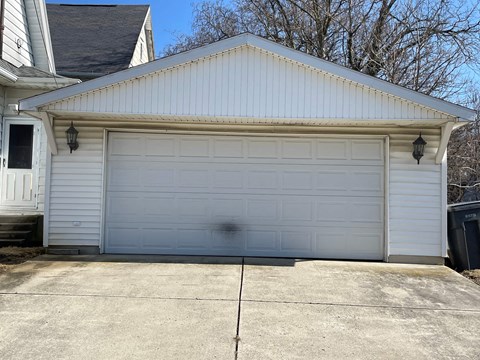 A white garage door is closed and there is a black trash can to the right of the garage.