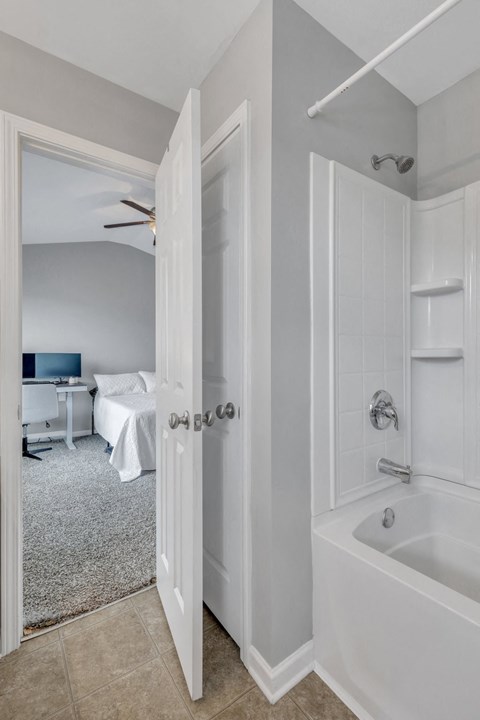 A white bathroom with a tub and a bedroom with a bed and a desk.