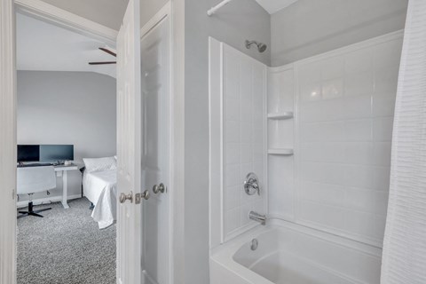 A white bathroom with a tub and a bedroom with a desk and chair.