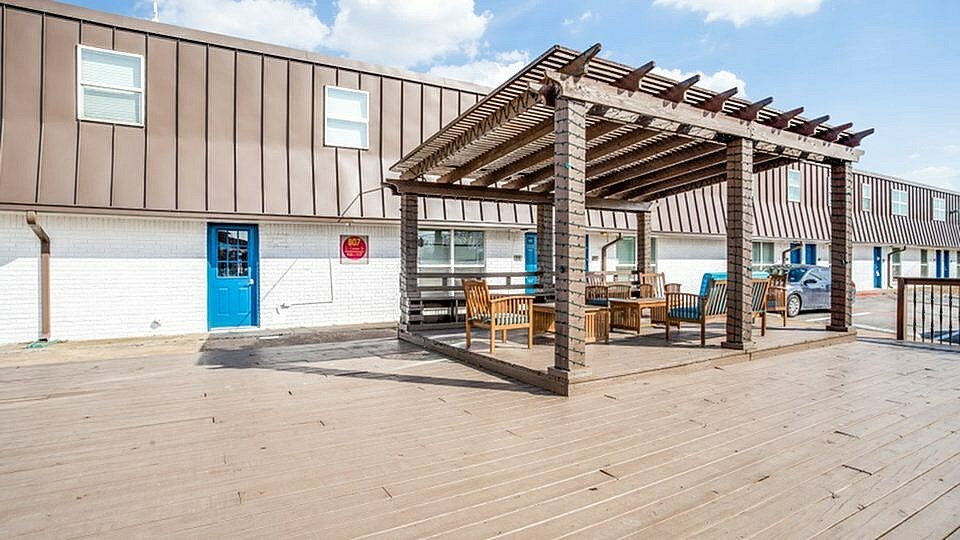 A wooden pergola with chairs and a table is in front of a building with a blue door.