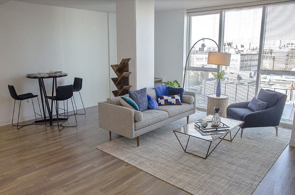 A living room with a grey couch, a black chair, a glass table and a wooden floor.