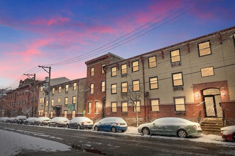 A row of cars are parked on a snowy street in front of apartment buildings.