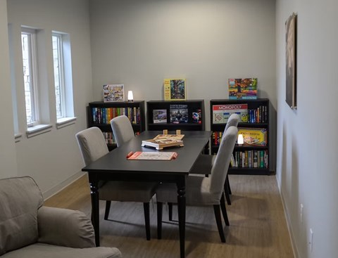 A black dining table with four grey chairs and a couch with a cushion in front of a bookshelf.