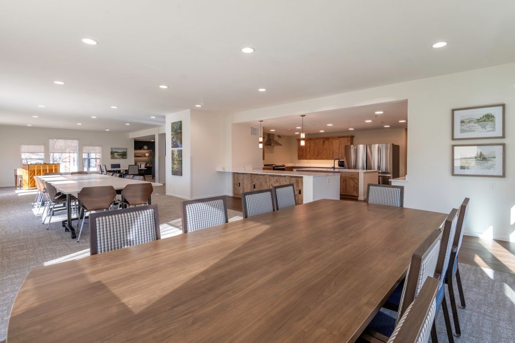 A large conference table with chairs is in the foreground of a well-lit room with a kitchen in the background.