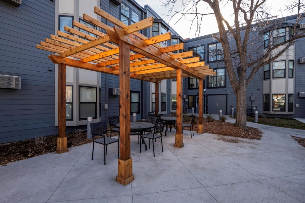 A wooden pergola with chairs and a table is in front of a building.