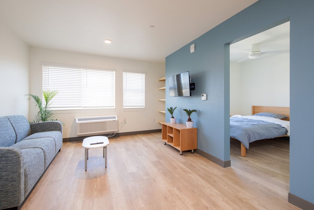 A living room with a grey couch, a white table, a window with blinds, a wooden shelf, and a bed in the background.