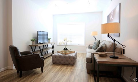 A living room with a brown chair, a grey ottoman, a wooden table, and a lamp.