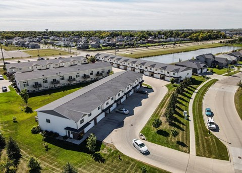 A large white building with a grey roof is surrounded by a grassy area and a road with cars.