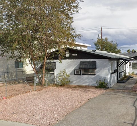 A white building with a black awning is surrounded by a gravel area and a tree.