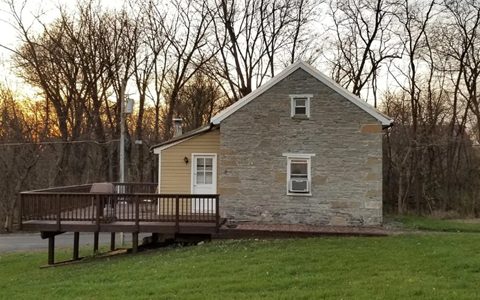 A small house with a porch and a deck is surrounded by trees.