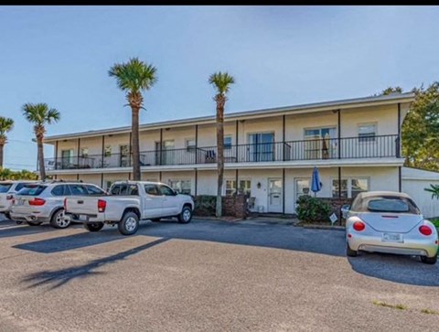 A white car is parked in front of a building with a balcony and palm trees.
