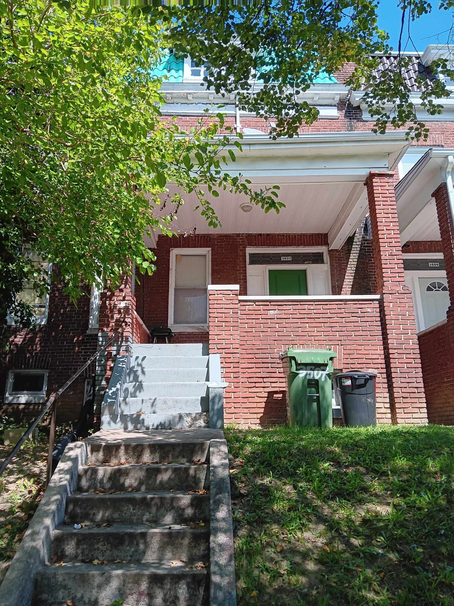 A red brick house with a green trash can and a tree in front.