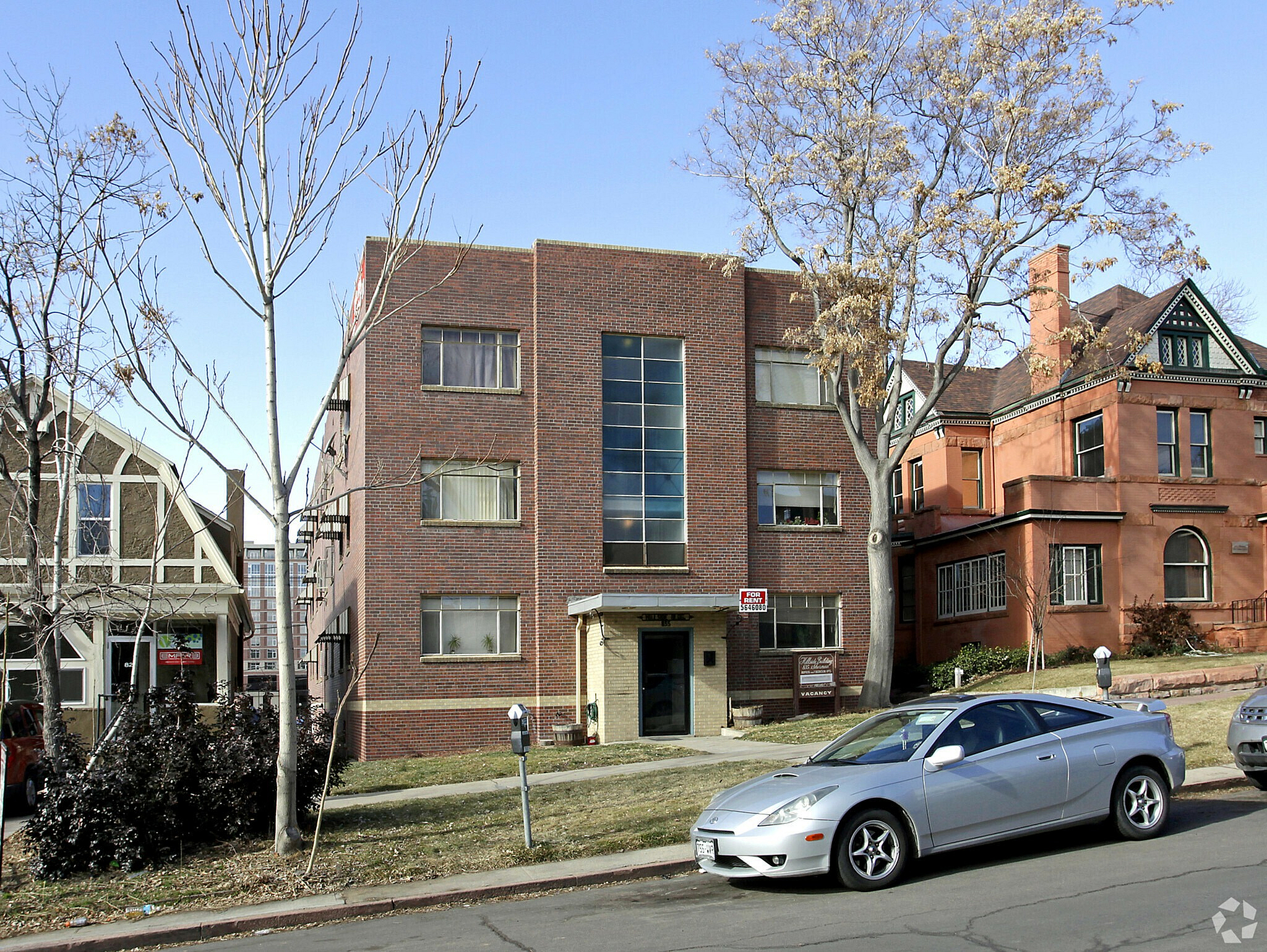A silver car is parked on the street in front of a brick building.